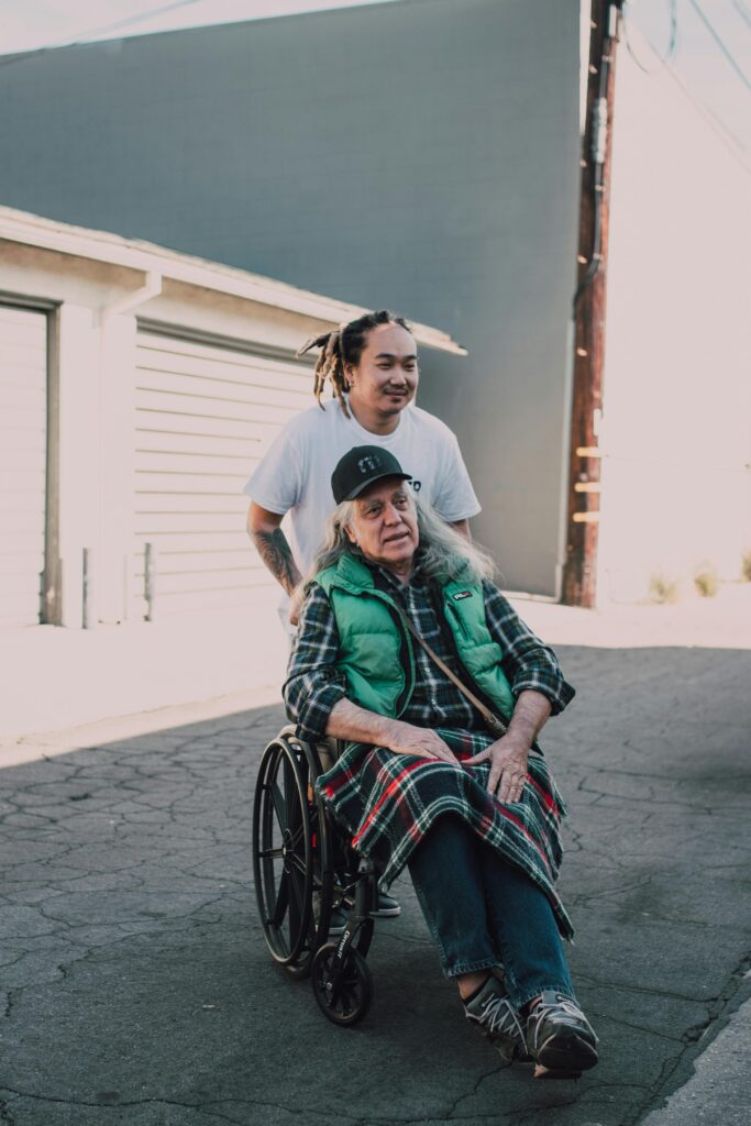 pexels-photo-6646919-6646919 A helpful young adult pushes a senior in a wheelchair on a sunny day, promoting kindness.